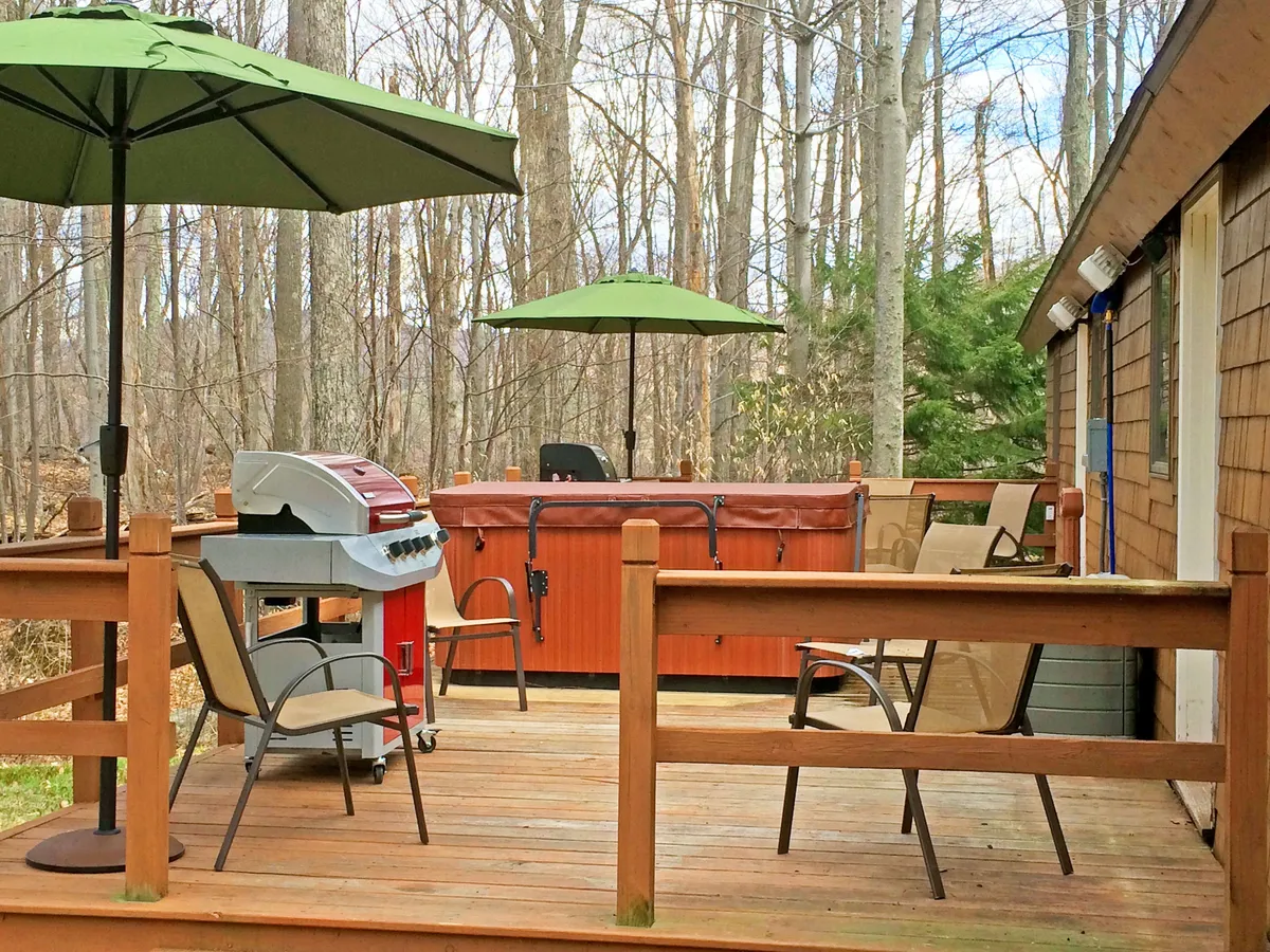 An inviting deck scene featuring a hot tub, grill, deck chairs, and umbrellas. The setup suggests a social space for outdoor dining, relaxation, and conversation, surrounded by natural scenery.