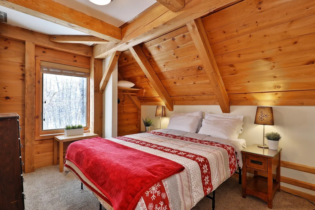 This image showcases a cozy bed with a warm red blanket situated beneath exposed wooden beams. A large window frames lush greenery just outside, bathing the room in natural light and presenting a direct visual connection to the natural surroundings. The inviting setup suggests both comfort and a strong link to the peaceful environment outdoors, making it an ideal setting for a remote worker seeking tranquility.