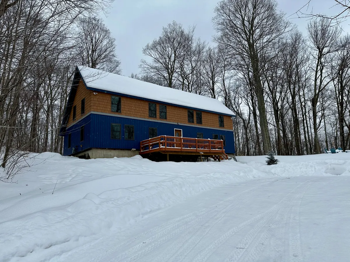 A two-tone colored duplex is shown in a snow-covered landscape, with a wooden deck extending from one side. The building clearly shows two separate units, highlighting the duplex layout. Snow blankets the ground and trees, evoking a sense of winter tranquility and privacy. The deck hints at shared or private outdoor gathering spaces for guests.