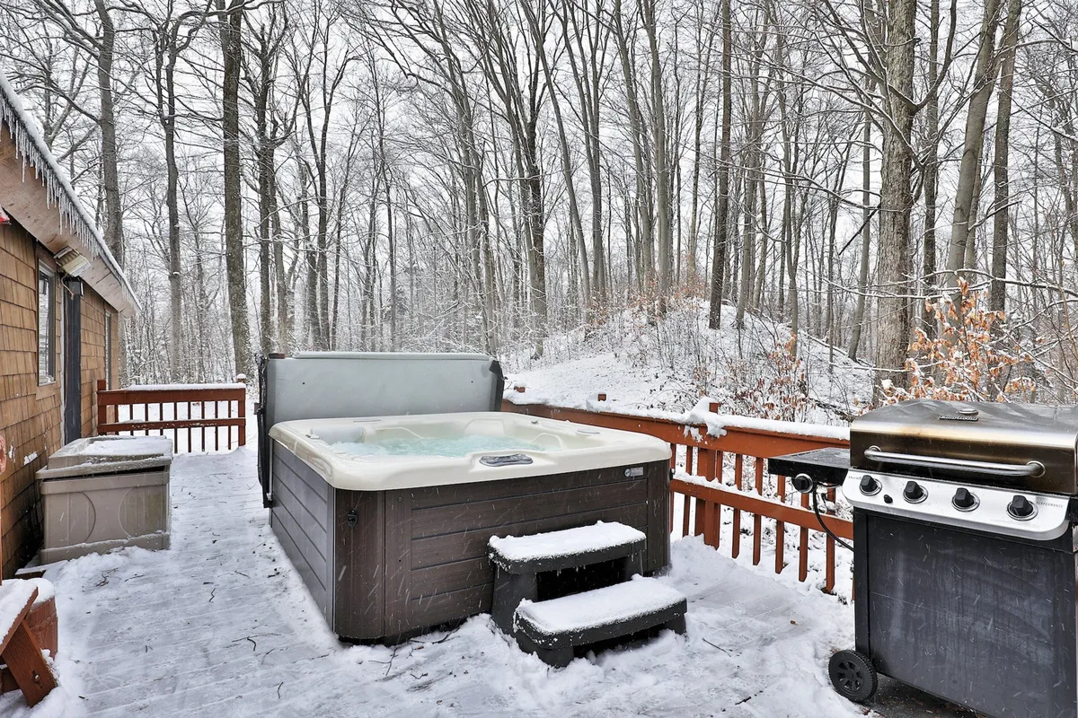 This exterior image showcases a bubbling hot tub and grill set against a tranquil backdrop of snow-dusted trees. The inviting steam from the tub is visible, and the scene hints at a cozy winter setting perfect for unwinding after outdoor activities.