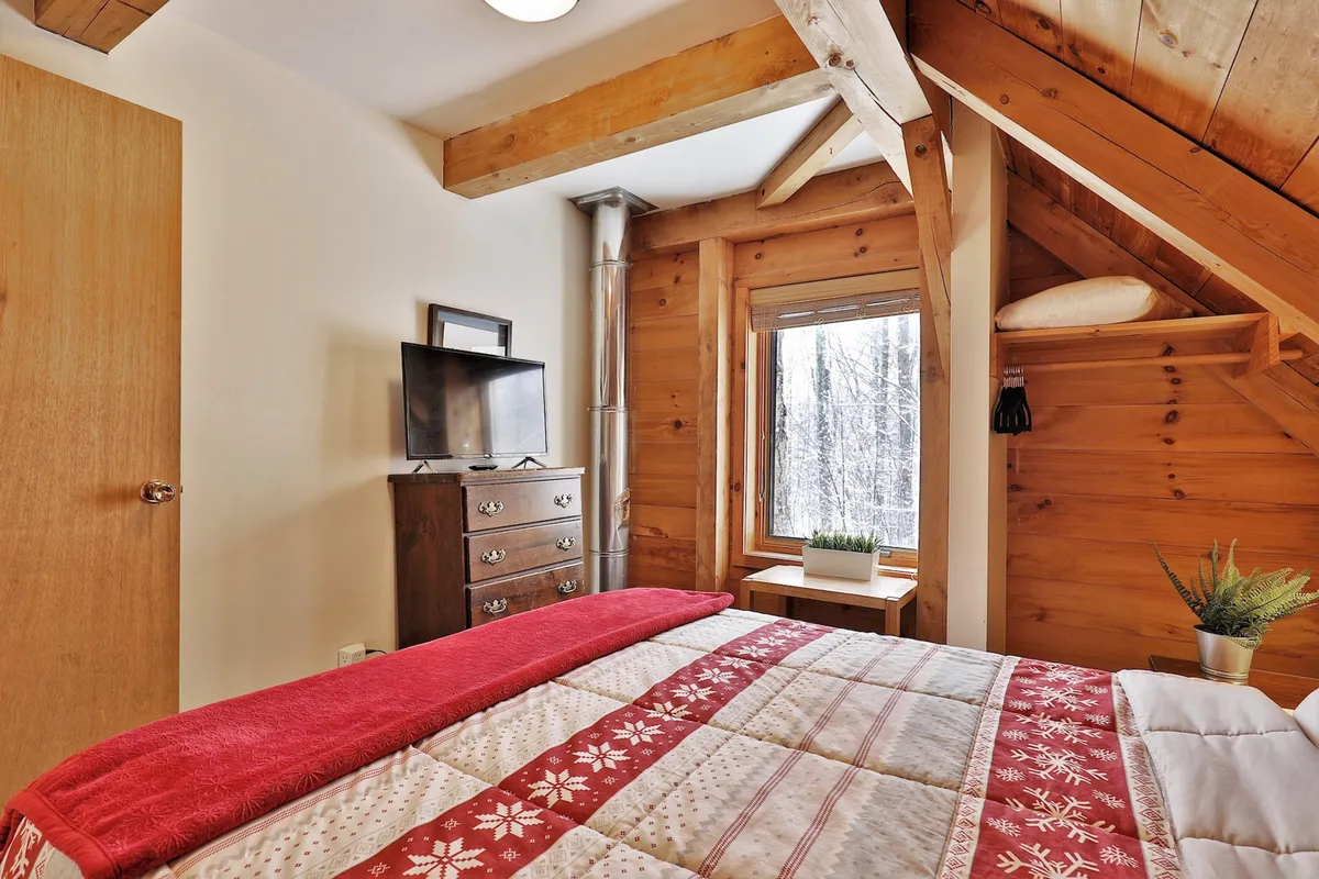 This image features a warm and inviting bedroom with a bed covered in a red quilt, rustic wooden furniture, and a window overlooking a snowy landscape. The setting is both cozy and inspiring, with the window view reinforcing the property's mountain setting and sense of seclusion, perfect for moments of relaxation or focused work.