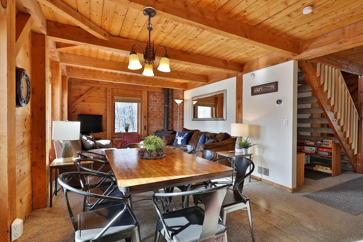 Inside, there’s a spacious open-plan area with a dining table, cozy living area, large window filling the room with light, and a staircase leading upstairs. The layout encourages communal activities like meals and game nights, while also offering space for relaxation.