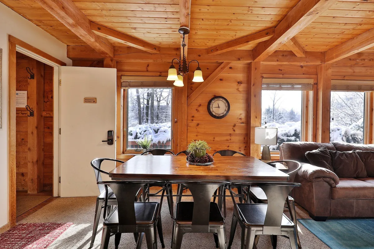 A warm, inviting interior scene featuring a large wooden dining table with black chairs and a comfortable sofa near expansive windows that reveal a snowy landscape. The space is ideal for group meals, game nights, or cozy conversations after a day outdoors.
