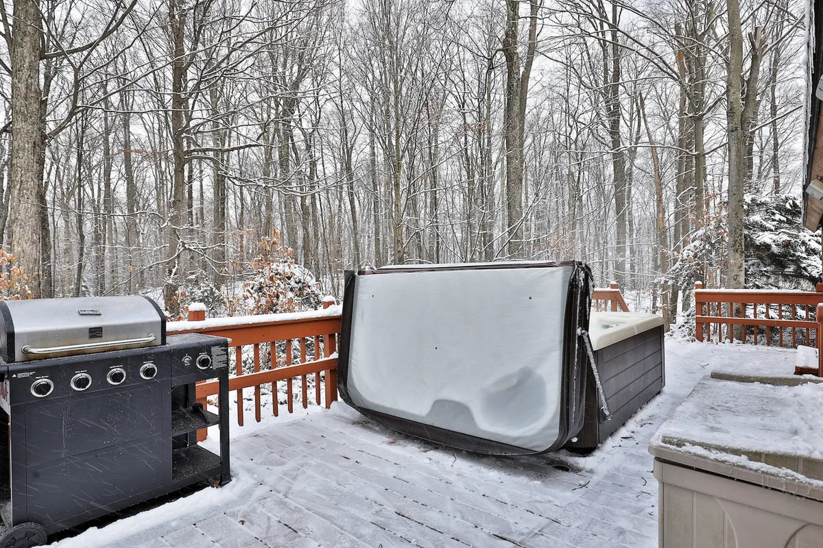 An exterior view featuring a grill and hot tub, surrounded by trees in the background. The outdoor area invites relaxation and social activities, with ample room for people and pets to enjoy nature year-round.