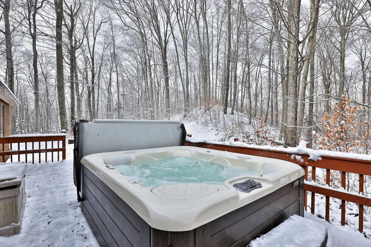 A serene winter scene showcasing a snow-framed wooden deck with a hot tub, surrounded by snow-covered trees. The intimate setting evokes feelings of warmth and relaxation even in colder months.