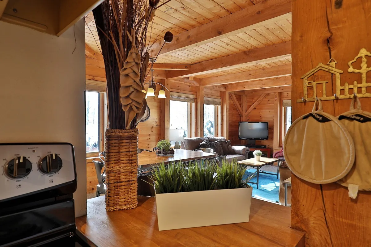 A welcoming great room features exposed wooden beams, large windows, comfortable seating, and a modern kitchen area. The space is designed for family gatherings, socializing, and enjoying the natural light, with plenty of room to relax or entertain.