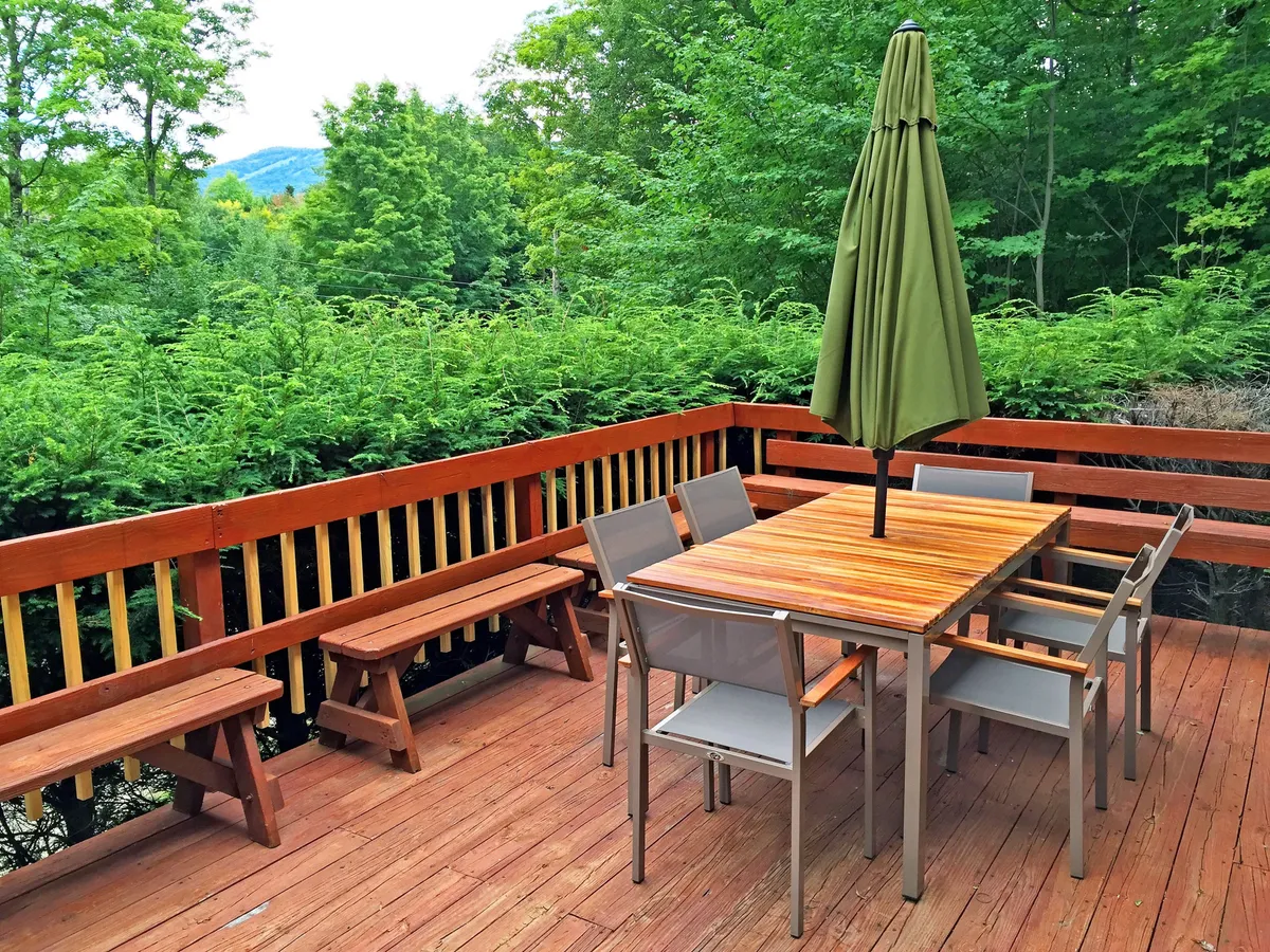 A wooden outdoor dining table with a green umbrella and benches, set amongst vibrant greenery. The space is ideal for outdoor meals or simply relaxing in nature with family, friends, or furry companions.