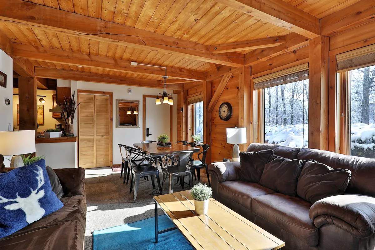 A cozy, sunlit common area showcasing wooden beams overhead, a spacious dining table, comfortable sofas, and large windows revealing scenic outdoor views. The design harmoniously blends rustic textures with an airy, inviting atmosphere, ideal for group meals, board games, or unwinding after outdoor adventures.