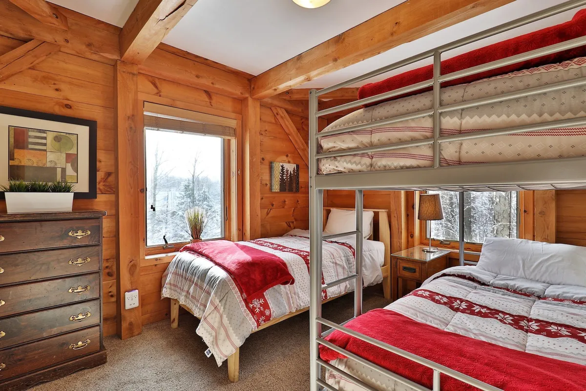 This image features a warm and inviting bedroom with a prominent bunk bed and a double bed, complemented by a chest of drawers and a window offering a snowy view. The arrangement hints at comfortable bedding and playful, family-friendly sleeping arrangements, ideal for both kids and adults.