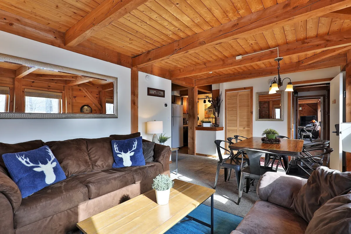 A warm, brightly lit interior featuring plush brown sofas with decorative deer pillows, a sturdy wooden dining table, and a view into the fully equipped kitchen. Modern touches like visible appliances and an open layout invite relaxation and social interaction, while the décor combines rustic charm with contemporary convenience.