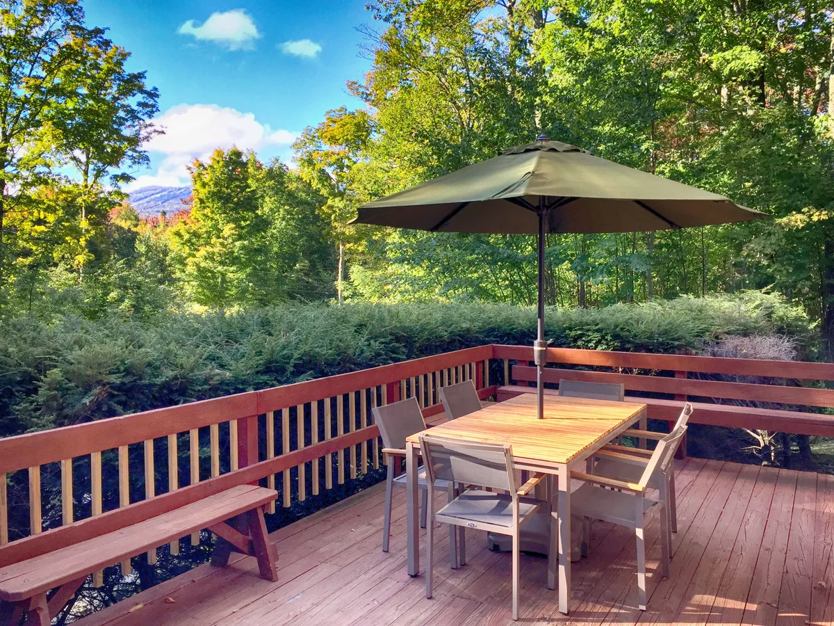 The outdoor dining area features a wooden table with an umbrella and seating, surrounded by greenery and a wooden railing. The setup is ideal for meals with family or friends, connecting the indoor and outdoor living experience.
