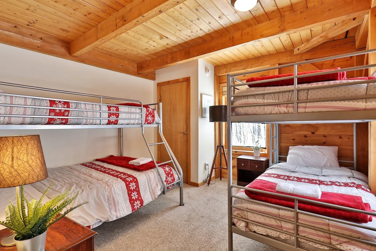 This interior shot showcases a bedroom with classic bunk beds, a single bed, a softly glowing lamp, and a window that lets in morning sunlight. The furnishings are arranged to evoke a sense of warmth and shared experiences, ideal for group or family stays.