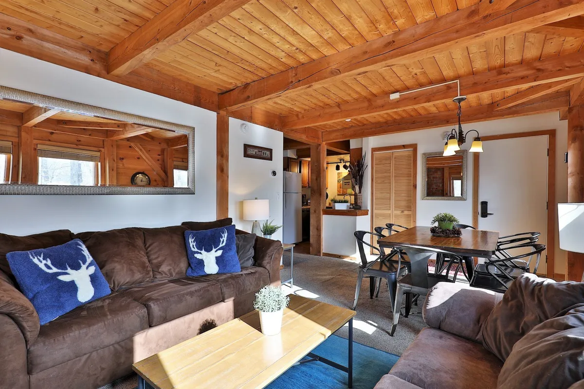 This image captures an open-concept living, dining, and kitchen area featuring exposed wooden beams, a sofa adorned with deer-themed pillows, a family-sized dining table, and a visible kitchen with modern amenities. Warm natural light pours in through the windows, highlighting the inviting, mountain-inspired décor and the seamless flow between social spaces.